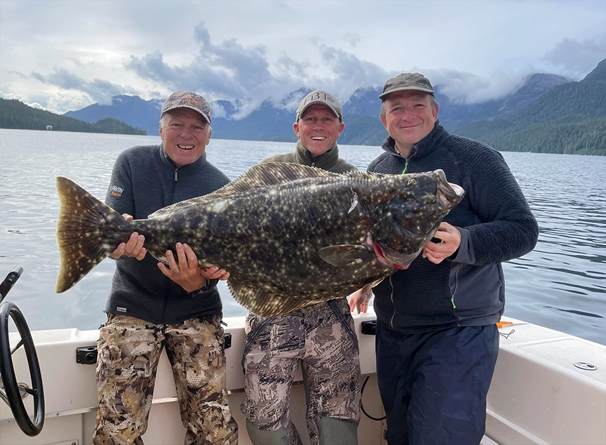 Two smiling guests holding up a massive halibut on a charter fishing trip with scenic coastal mountains in the background