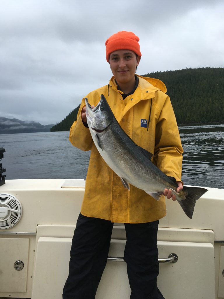 Smiling fisherman in a yellow rain jacket and orange toque holding a freshly caught salmon on a boat in Kitimat waters