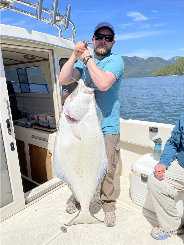Angler standing on a boat holding a massive Pacific halibut with mountains and forest in the background